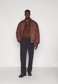 Braune Steppjacke mit Reißverschluss, strukturierter brauner Pullover, schwarze Hose und schwarze Sneaker. Einfaches Design, schlanker Schnitt und keine sichtbaren Logos.