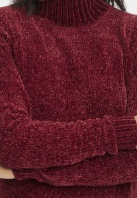 Pull en tricot bordeaux avec un col montant côtelé, présentant une surface texturée et duveteuse ainsi que des poignets ajustés. Le tissu semble épais et confortable.