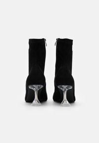 Botas de tobillo de ante negro con cierre de cremallera, que presentan un tacón de cristal transparente y un diseño de punta afilada. Textura suave y silueta elegante.