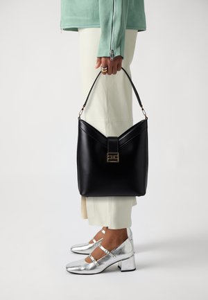 Borsa in pelle nera con un design a V, hardware dorato e chiusura. Indossata con tacchi metallici argentati e pantaloni chiari.