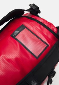 Red backpack made of textured material, featuring a hexagonal-patterned accent, adjustable black straps, and a logo patch.
