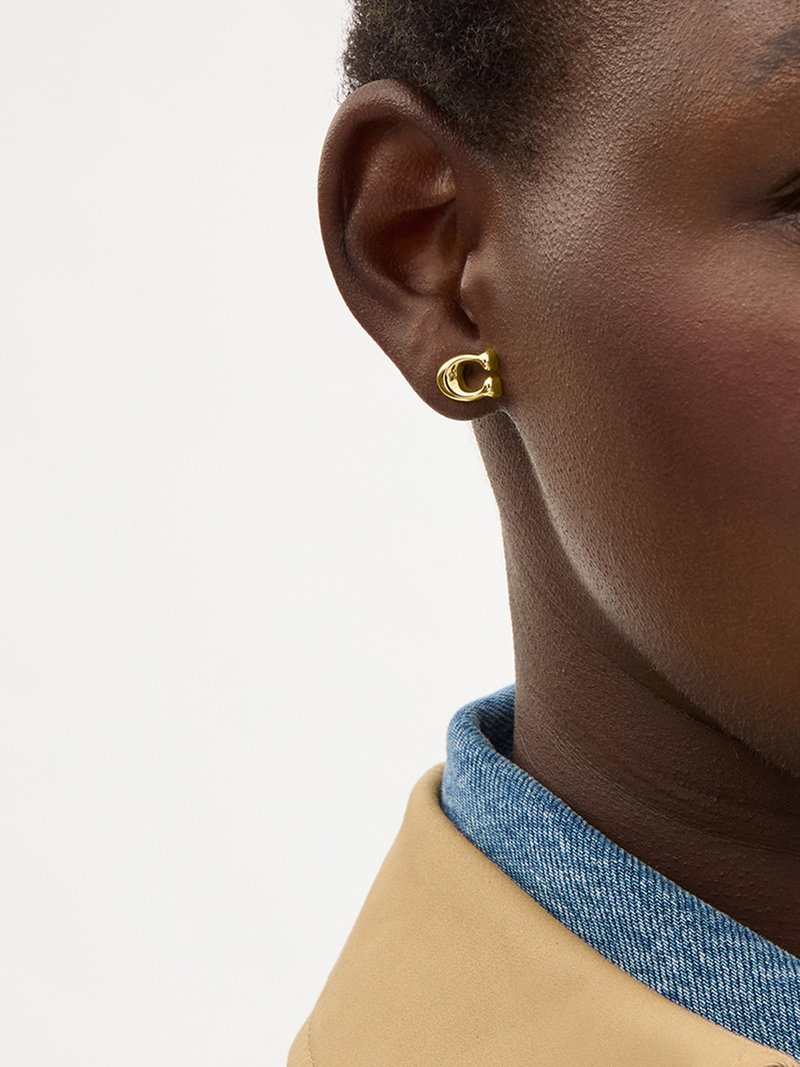 Nahaufnahme eines Ohrs einer Person, die einen kleinen goldenen Creolen-Ohrring in Form des Buchstabens C trägt, mit einer beigefarbenen Jacke und einem blauen Kragen im Hintergrund sichtbar.