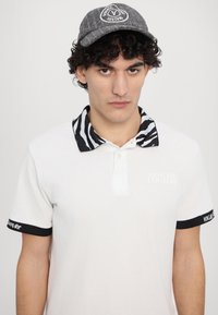 Camisa polo branca com colarinho padrão zebra preto e branco, detalhes pretos nas mangas, apresentando a marca "VERSACE JEANS COUTURE". Boné cinza na cabeça.