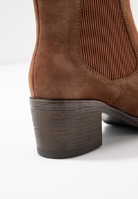 Brown suede ankle boot with ribbed elastic side, rounded toe, and a textured brown stacked heel. Smooth finish with subtle stitching details.