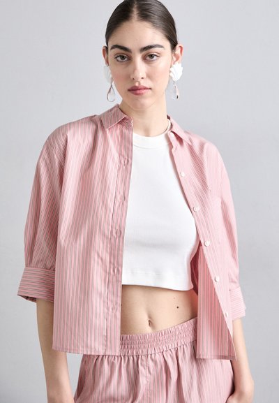 Pink striped shirt with a button-up design and rolled sleeves, worn over a white cropped ribbed top. Smooth texture, casual fit.