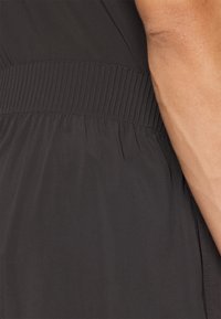 Svart plaggdetalj med en samlet, elastisk midje med vertikale folder, jevn tekstur og myk stoffoverflate.