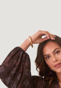Brazalete de metal dorado llevado en una muñeca, redondo y suave con un acabado pulido, en contraste con una blusa marrón de estampado con mangas abullonadas.