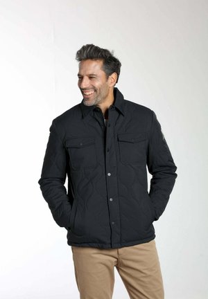Homme souriant portant une veste matelassée noire avec des poches frontales et un pantalon beige, les mains dans les poches, sur un fond clair uni.