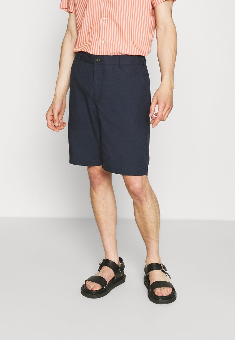 Man med marinblå shorts, svarta spännremssandaler och en kortärmad rosa och vit randig skjorta, stående på ett ljusgrått golv.