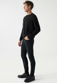 Suéter negro de punto con cuello redondo, combinado con jeans negros ajustados. El calzado presenta suelas gruesas y cordones, todo sobre un fondo blanco liso.