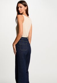 Beige sleeveless top paired with high-waisted dark blue denim pants; features classic stitching and back pockets. Smooth fabric texture.