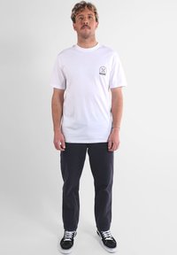 T-shirt en coton blanc avec un col rond et un petit logo imprimé sur le côté gauche de la poitrine ; assorti à un pantalon foncé et des baskets noires.