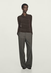 Woman stands with hands in pockets wearing a fitted brown long-sleeve wrap top and loose gray wide-leg trousers against a plain background.