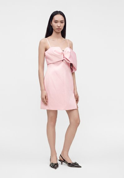 Jeune femme portant une mini-robe bustier rose pâle avec un grand nœud et des escarpins slingback métalliques à bout pointu.