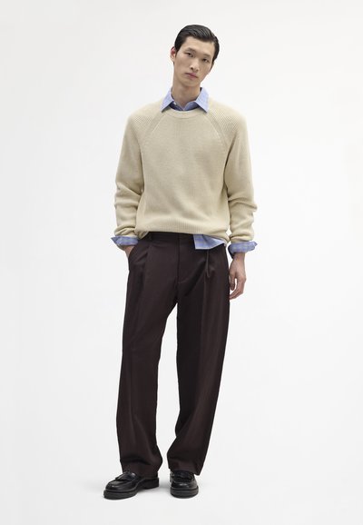 Bézs kötött pulóver raglán ujjakkal világoskék kockás ing felett, sötétbarna, széles szárú nadrággal és fekete cipővel párosítva.