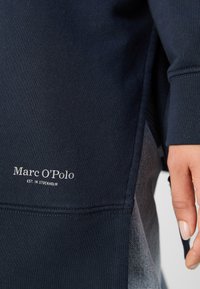 Tissu bleu foncé avec logo blanc Marc O'Polo et texte "Est. in Stockholm", montrant la main d'une personne et une partie de la manche.