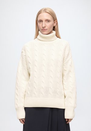 Femme aux cheveux blonds raides portant un pull blanc à col roulé en maille torsadée et une jupe sombre sur un fond uni.