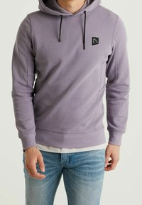 Hoodie violet en tissu doux, doté d'une capuche avec cordon de serrage, de poignets côtelés et d'un bas côtelé, ainsi que d'un petit patch logo noir sur la poitrine.