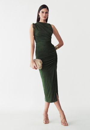 Kvinde iført en ærmeløs mørkegrøn midikjole med rynkede detaljer og en sideslids, med en vævet clutch i hånden og iført gyldne sandaler og smykker.