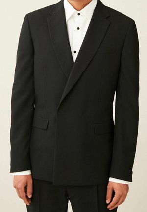 Homme portant une veste de costume noire sur une chemise blanche avec des boutons noirs, debout devant un fond uni.