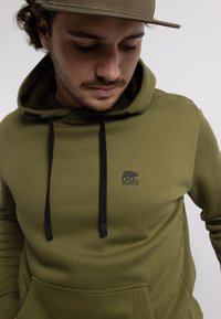 Olivenfarbener Kapuzenpullover aus weichem Stoff mit einer Kängurutasche, schwarzen Kordeln und einem kleinen Logo, das einen Bären und das Wort "WORKS" zeigt.
