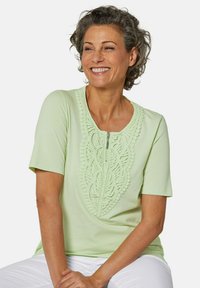 Top verde chiaro a maniche corte con un design frontale testurizzato e ricamato, dotato di zip. Tessuto morbido e vestibilità comoda, abbinato a pantaloni bianchi.