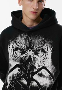 Mladý človek v čiernej mikine s kapucňou, na ktorej sa nachádza opotrebovaný biely grafický motív intenzívnej lebky s bleskovými detailmi na prednej strane.