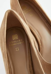Béžové semišové lodičky na vysokém podpatku se zaoblenou špičkou. Zlaté logo "Forever Comfort" na stélce. Hladký vnitřní podšívkový materiál, měkká textura.