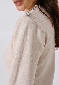 Maglione beige con un lavorato a trama. Presenta bottoni decorativi argento sulla spalla, di forma rotonda con un bordo intrecciato.