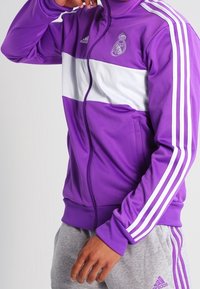 Fialová zipová bunda s bílým vodorovným pruhem, která má logo Real Madrid a tři bílé pruhy Adidas na rukávech.