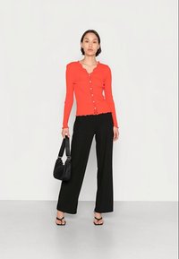 Cardigan vermelho de manga longa com bainha franzida e botões, combinado com calças pretas de perna larga e uma mala preta. Modelo a usar sandálias com tiras.