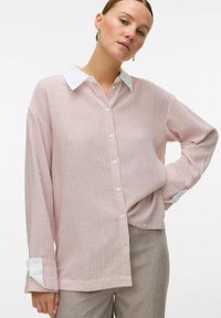 Button-up skjorta med vertikala röda och vita ränder, långa ärmar, vit krage och uppvikta manschetter. Tillverkad av lättviktsmaterial.
