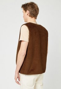 Brauner Cord-Weste mit vertikaler Rippstruktur, versehen mit weiten Armausschnitten und einem abgerundeten Saum, getragen über einem cremefarbenen T-Shirt.