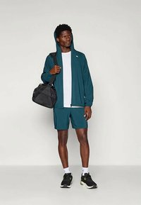 Türkiis värvi kergest jakist kapuutsiga, valgest ümmarguse kaelusega särgist, sobivatest türkiissinistest lühikestest pükstest ja mustadest sporditossudest. Hoides musta spordikotti.