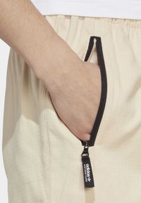 Pantalones beige texturizados con un bolsillo con cremallera visible. Cremallera negra con un tirador de marca. Mano descansando dentro del bolsillo.