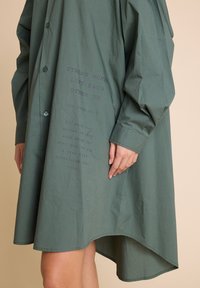 Grønn bomullskjole med lange ermer, knappelukking og asymmetrisk kant. Har trykt tekstdesign på forsiden.