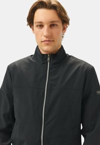 Giacca nera con zip e colletto alto, dalla texture liscia. Presenta una zip silver e un logo sulla manica sinistra. Vestibilità aderente.