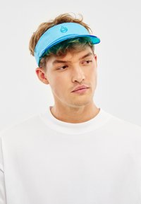 Mladý muž s krátkymi kučeravými vlasmi má na sebe bielu košeľu a modrú šiltovku, pozerá sa mierne doprava na jednofarebnom pozadí.