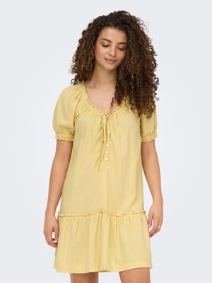 Femme aux cheveux bouclés portant une robe jaune courte avec des manches bouffantes et des détails de boutons, debout devant un fond clair uni.