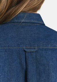 Back view of person with light brown hair wearing a blue denim jacket featuring yellow stitching and a back yoke detail.
