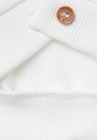 Tejido de suéter blanco con textura acanalada. Presenta un botón redondo marrón con logotipo grabado. Forro interior suave y liso visible.