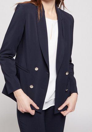 Blazer bleu marine à double boutonnage avec des boutons argentés, revers cranté et poches à rabat, porté sur un top blanc à rayures verticales.
