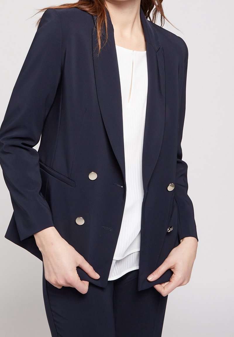 Blazer doppiopetto blu navy con bottoni argentati, revers a scollo, e tasche a pattina, indossato su una maglietta bianca con righe verticali.