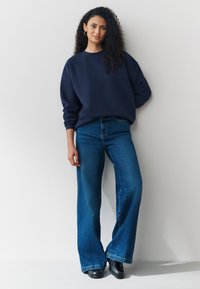 Tamnoplava majica s širokim rukavima uparena s visokim strukom, zvonastim trapericama srednje plave boje. Model nosi crne cipele i stoji na svijetloj pozadini.