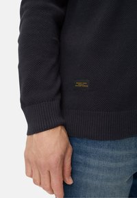 Dunkelblauer Strickpullover mit strukturiertem Muster und gerippten Bündchen. Verfügt über ein kleines rechteckiges Etikett mit goldener Schrift nahe dem Saum.