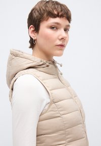 Μπεζ φουσκωτό γιλέκο με κουκούλα, με οριζόντια ραφή και εμπρόσθιο φερμουάρ. Φορεμένο πάνω από λευκό μακρυμάνικο πουκάμισο.