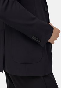 Černý upravený blazer s hladkou texturou látky, slim-fit designem, se čtyřmi knoflíkovými detaily na manžetě a viditelnými kapsami.