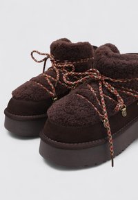 Botas de plataforma de ante marrón con partes superiores de forro polar, con cordones tejidos de colores y detalles metálicos. Superficies suaves y texturizadas visibles.