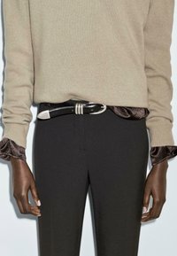 Maglione beige con texture a coste, indossato sopra una blusa nera in seta. Cintura nera con accenti argentati e forma elegante. Pantaloni neri attillati.