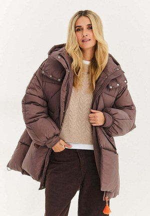 Brązowa puchowa kurtka z kapturem, z zatrzaskami i pomarańczowym suwakiem. Noszona na beżowym swetrze w warkocze i ciemnych spodniach.